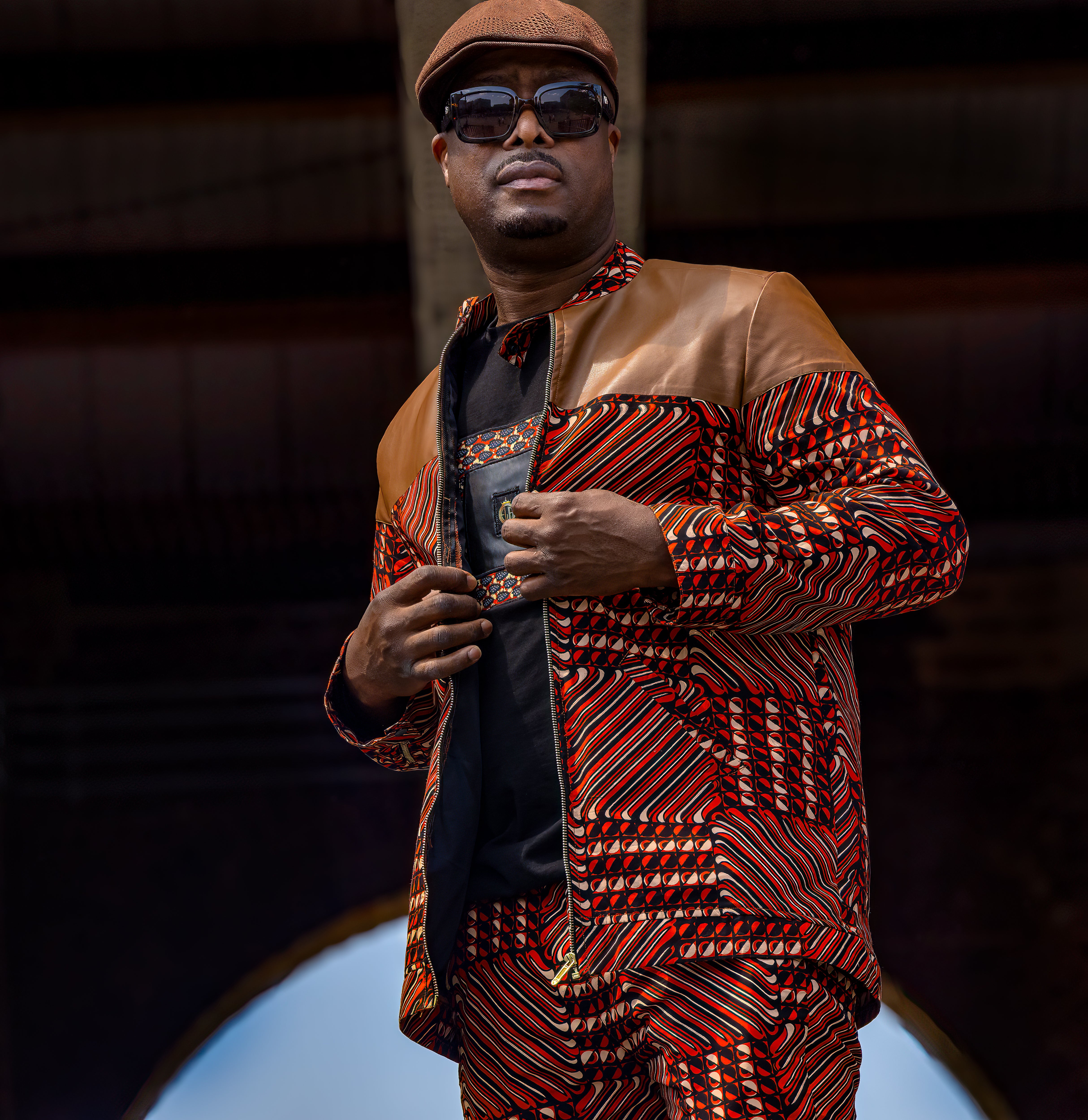 Man wearing a patterned African print jacket and sunglasses against a dark background
